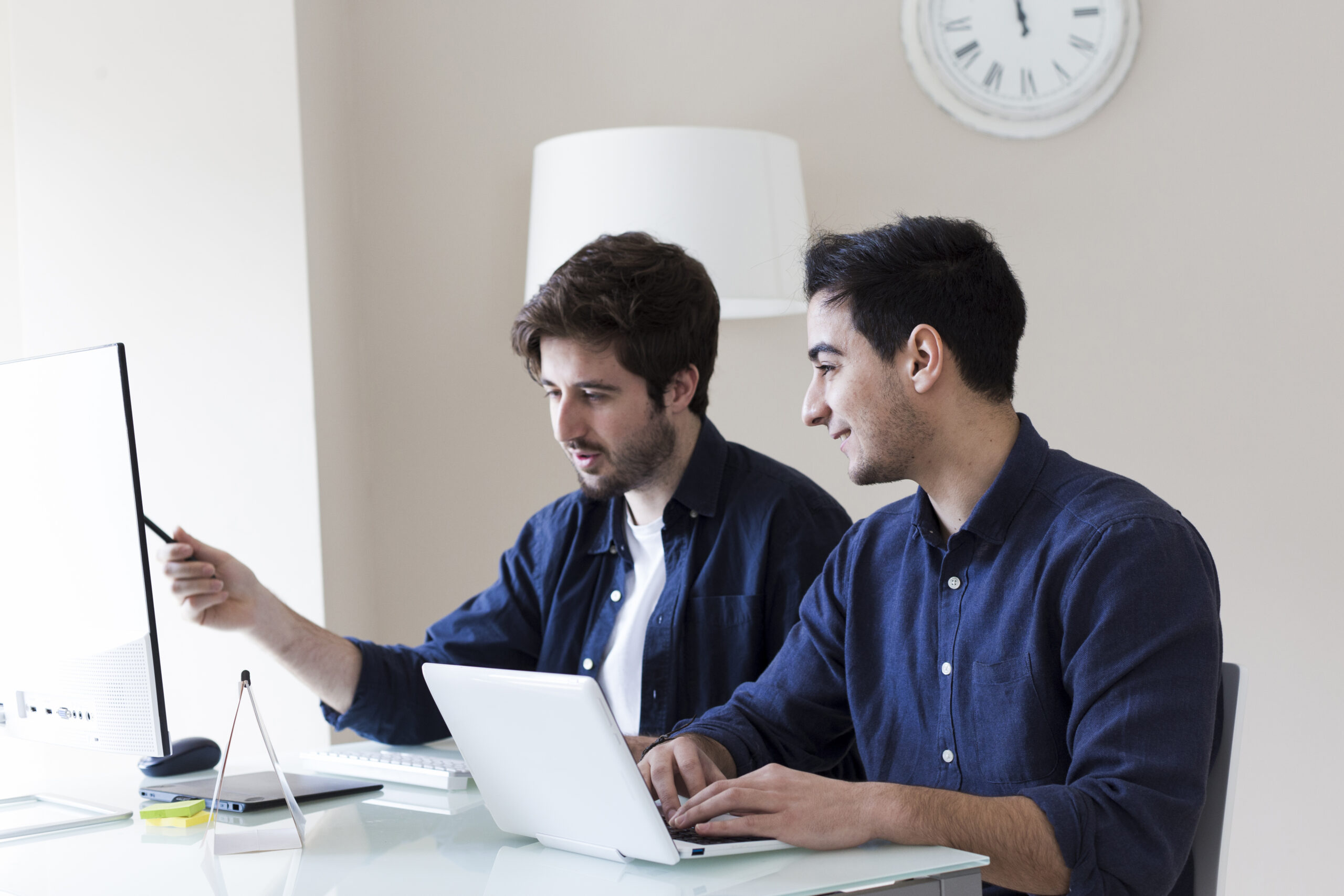 Two colleagues discussing a work project at a desk, one pointing at a computer screen while the other uses a laptop.