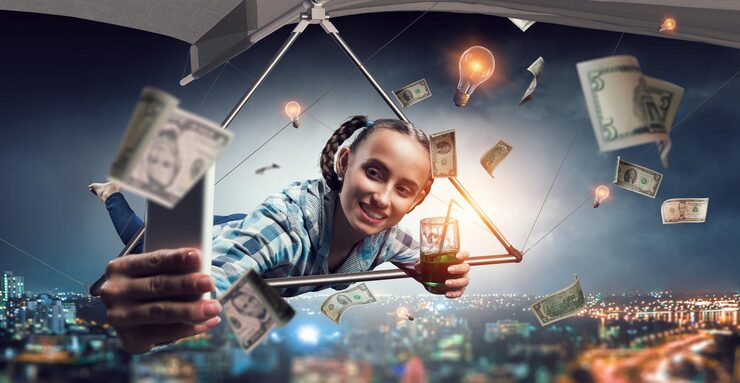 Khush mizaj larki hang glider par uḍti hui, mobile se selfie leti hui, shehar ke upar paison aur light bulbs ke sath.